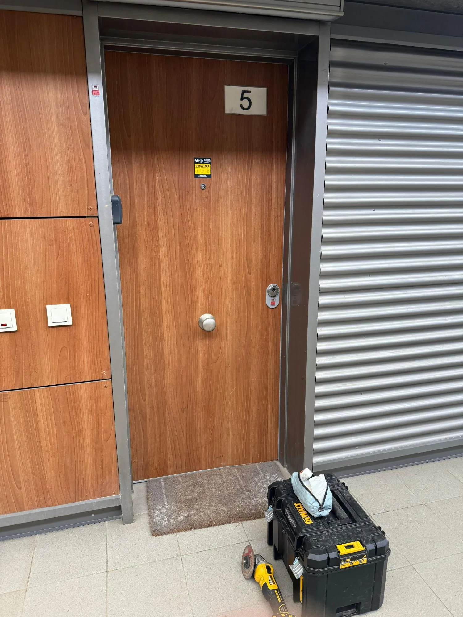 Puerta de madera de una vivienda en Pozuelo de Alarcón, con escudo de seguridad en el bombín y la caja de herramientas y la radial de nuestro técnico en el suelo.